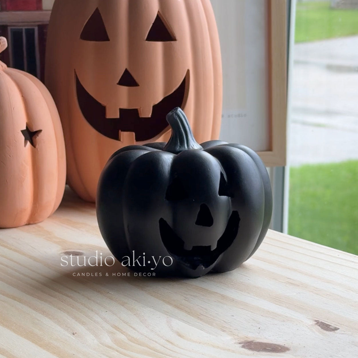 Black pumpkin-shaped candle on a wooden surface with orange pumpkins in the background.