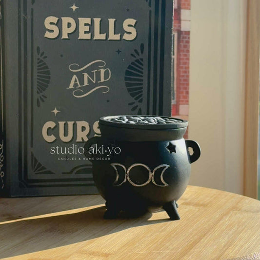 Black cauldron with candles on a wooden surface next to a book titled 'Spells and Curses' by studio akiyo.