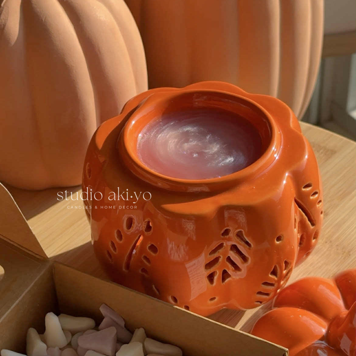 Orange ceramic wax warmer with a candle inside, surrounded by pumpkins and decorative items on a wooden surface.