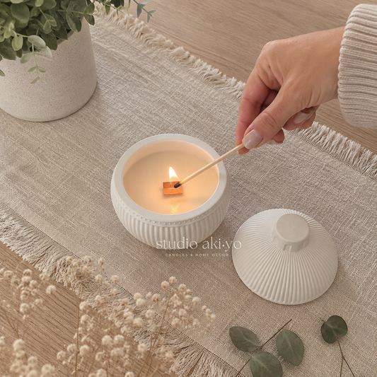 A white ribbed ceramic candle jar with its matching lid placed beside it on a beige woven runner. A hand is lighting a small wooden match, and the candle burns inside the open jar.