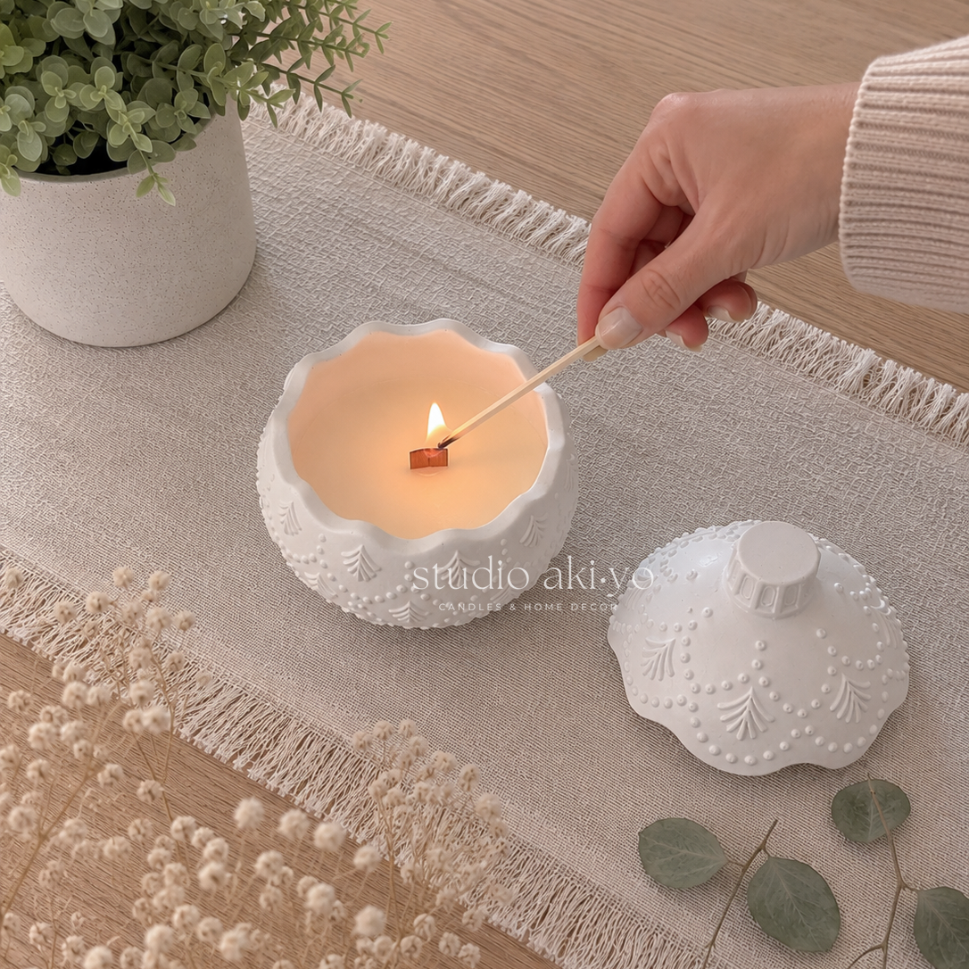 A white textured ceramic candle holder with a matching lid sits on a beige fringed runner. A hand is lighting a candle with a wooden match.