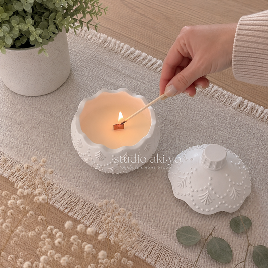 A white textured ceramic candle holder with a matching lid sits on a beige fringed runner. A hand is lighting a candle with a wooden match.