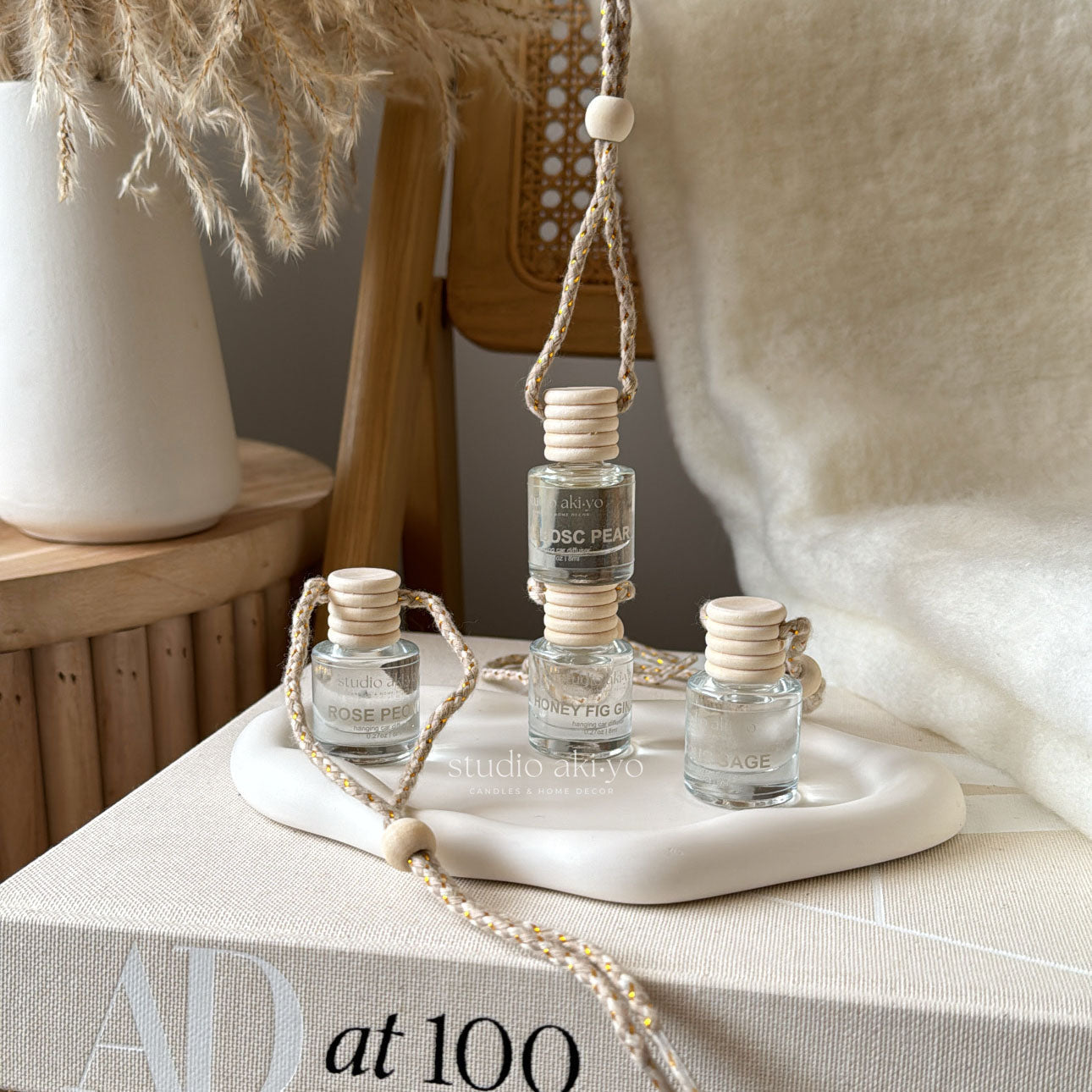 A hanging car diffuser with three small bottles of fragrance, suspended by a rope, displayed on a white surface, with a decorative setting in the background.