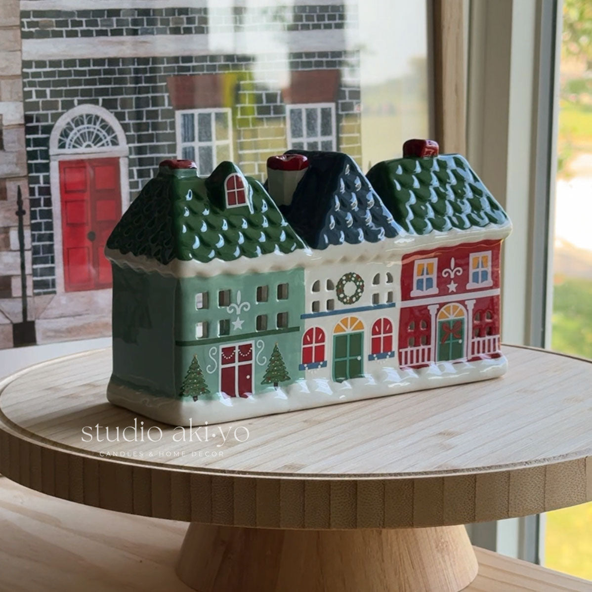 A ceramic Christmas village house figurine with three colorful houses (green, white, and red) on a wooden display stand in front of a window.