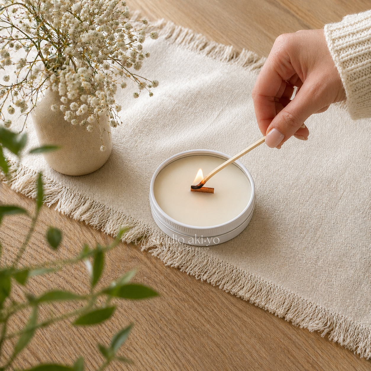 A round white candle tin on a beige fringed runner, with a hand lighting the candle using a wooden match. A small vase of baby's breath and green leaves are in the background on a wooden table.