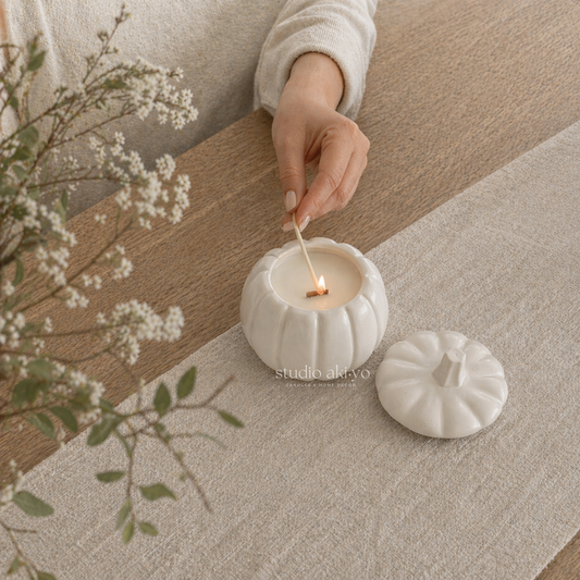 White ceramic candle holder with a lit candle on a wooden surface with white flowers.