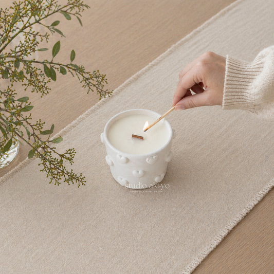 Hand lighting reverie wooden wick candle in a white ceramic holder on a wooden surface with a white cloth and greenery.