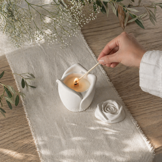 Hand lighting a candle in a white ceramic holder on a wooden surface with a white cloth and greenery.
