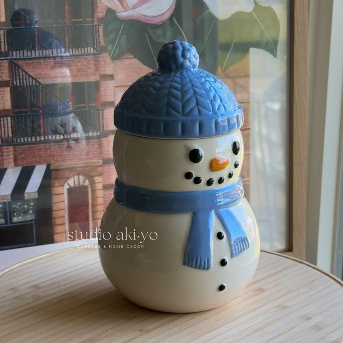 A ceramic snowman figurine with a blue knit hat and scarf, set on a light wooden table in front of a window with a cityscape reflection.