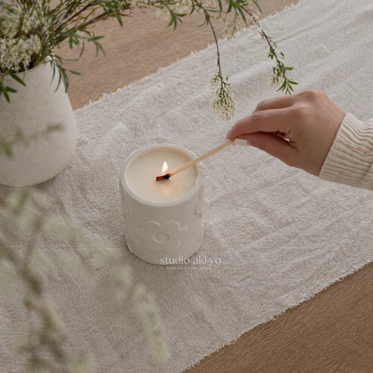 Hand lighting a candle in a white ceramic holder on a wooden surface with a white cloth and greenery.