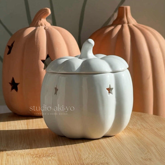 White pumpkin-shaped ceramic jar with a lid on a wooden surface, with two orange pumpkins in the background.