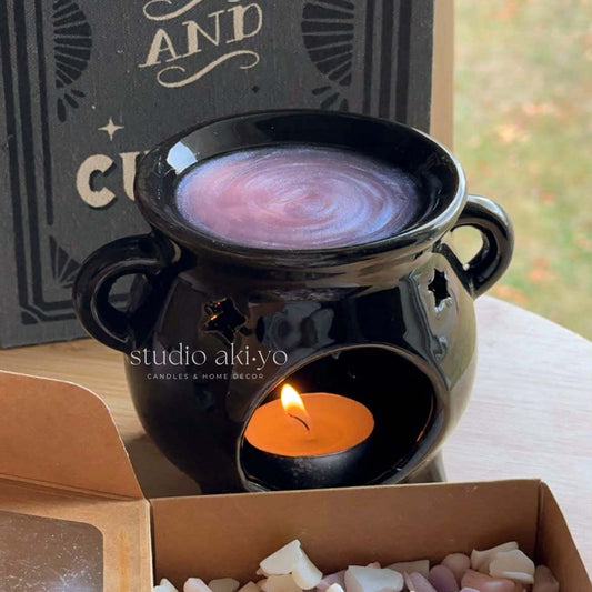 Black ceramic wtichy cauldron wax warmer with a lit candle inside, surrounded by decorative items on a table.