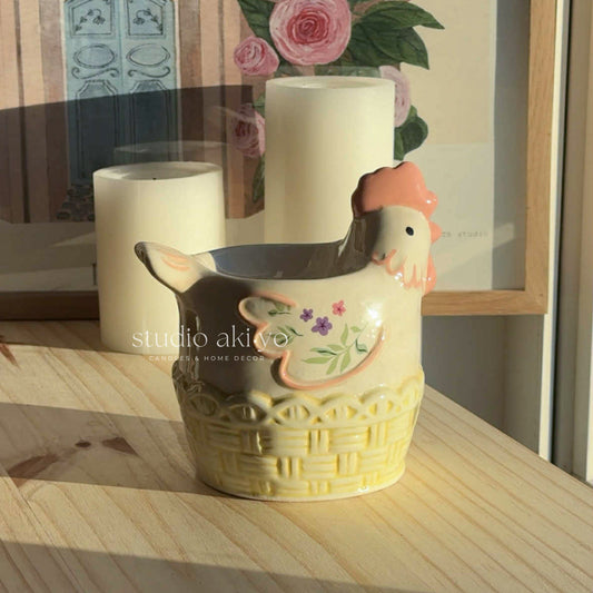 Decorative ceramic chicken-shaped vase on a wooden surface with a floral painting in the background.