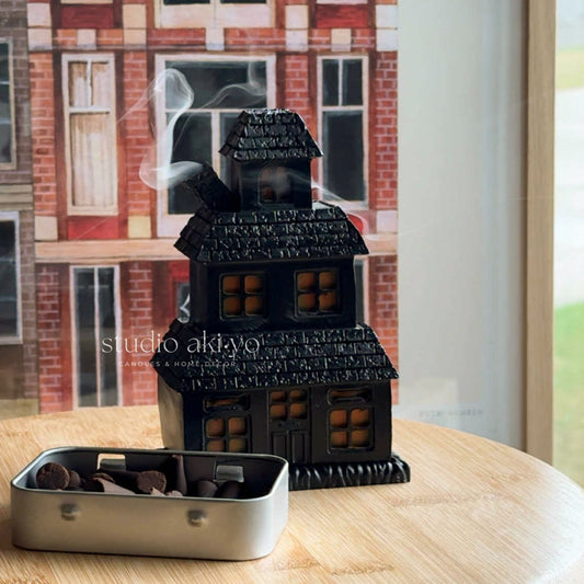 Black house-shaped incense burner with smoke on a wooden table, blurred building exterior in the background.