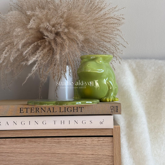 Green ceramic frog figurine on books with decorative pampas grass and a white blanket.