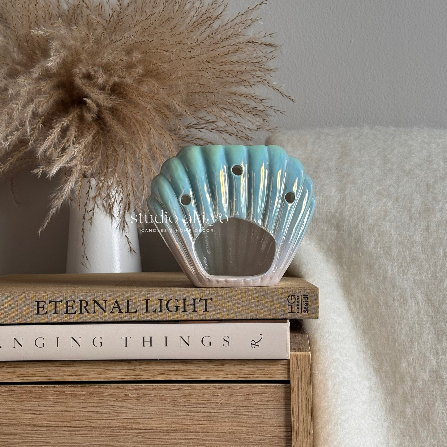 Decorative shell-shaped wax melt burner on books with a vase of dried pampas grass in the background.