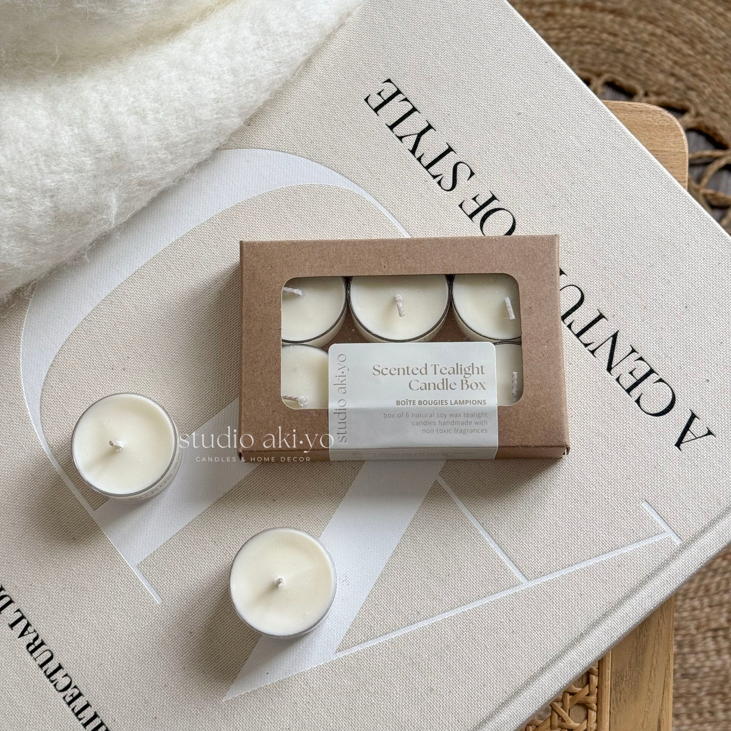 A box of six white tealight candles with a 'Studio Akiyo' label on it, displayed next to a lit candle on a white plate, with a white vase and decorative items in the background.