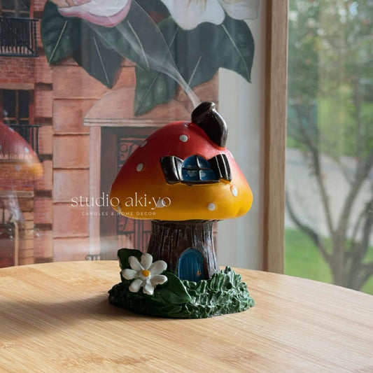 Decorative mushroom-shaped toadstool incense cone burner with a small door and flower on a wooden surface. Enjoy the smoke going through the little chimney for those whimsical cottagecore fairy vibes.