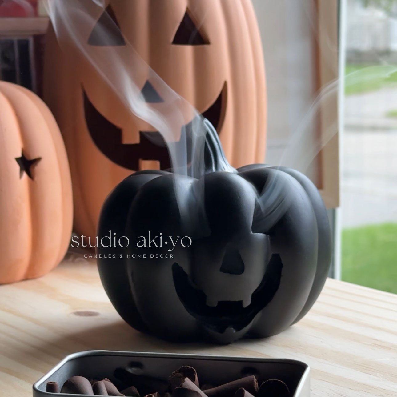 Black pumpkin-shaped diffuser with smoke emitting, surrounded by pumpkins, on a wooden surface.
