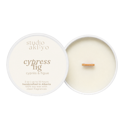 An open soy wax candle by studio akiyo, showing the cream-colored wax and wick inside a white round tin next to its lid, which is printed with the scent name 'cypress fig' and notes 'handcrafted in Alberta'.