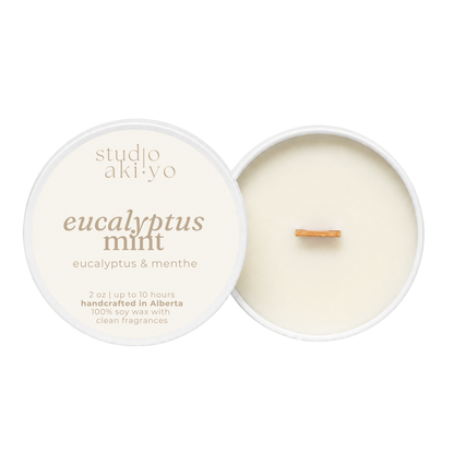 Studio Akiyo eucalyptus mint soy wax candle shown open with the cream-colored lid on the left and the candle vessel on the right, featuring a small wooden wick and a single eucalyptus leaf on the wax surface.