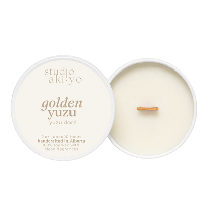 A soy wax candle shown open with its lid beside it on a white background. The lid is cream-colored with gold text reading 'studio aki·yo' and 'golden yuzu'.