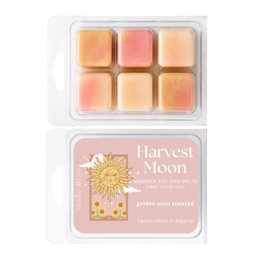 Two clear plastic clamshell packages of Studio Above Harvest Moon soy wax melts, showing six pastel wax cubes in peach, pink, and golden yellow tones, with the back label visible below.