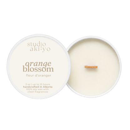 A soy wax candle by studio aki yo named 'orange blossom', shown with its cream-colored lid featuring gold text next to the open candle container displaying the creamy wax and a small wooden wick.