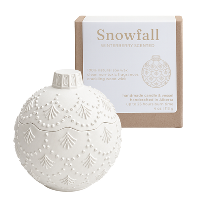 A white ornament-shaped scented candle with embossed tree and bead patterns sits in front of its kraft paper box. The box is labeled 'Snowfall' and 'Winterberry Scented.