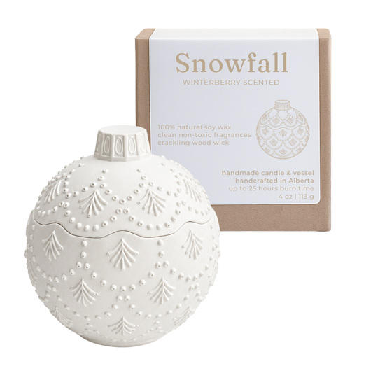 A white ornament-shaped scented candle with embossed tree and bead patterns sits in front of its kraft paper box. The box is labeled 'Snowfall' and 'Winterberry Scented.