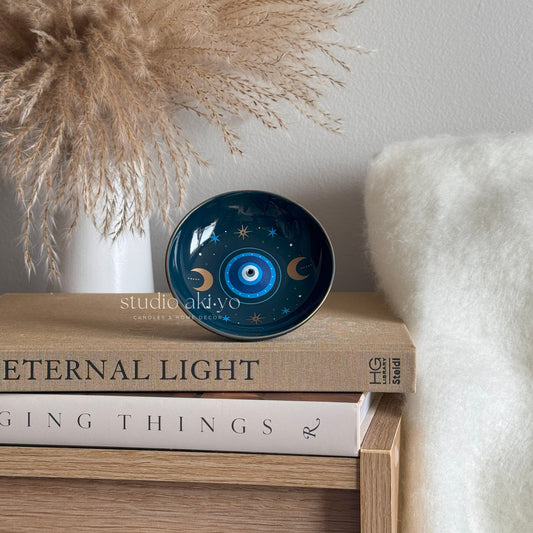 Blue all seeing eye incense stick holder on a stack of books, with pampas grass and a white vase in the background.