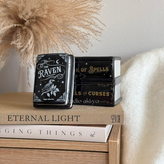 Two black books titled 'The Raven' and 'Book of Spells' on a wooden surface with decorative elements.