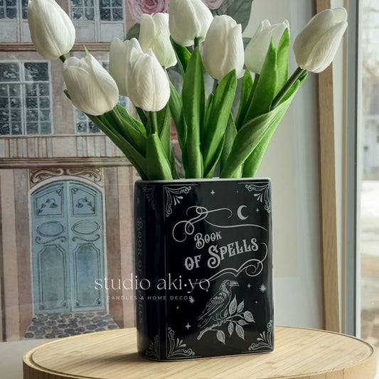 Black decorative vase with 'Book of Spells' design holding white tulips on a wooden surface.