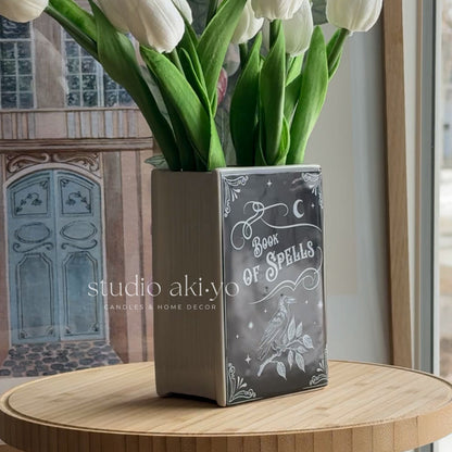 Decorative vase with 'Book of Spells' design on a wooden surface, featuring white tulips.
