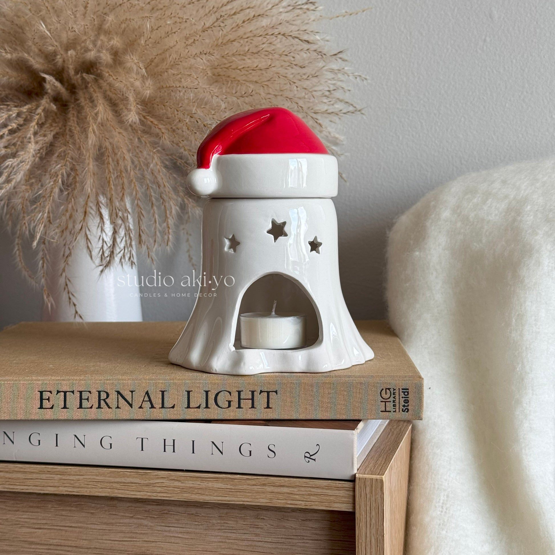 White ceramic candle holder with a red top on books, next to decorative pampas grass and a white blanket.