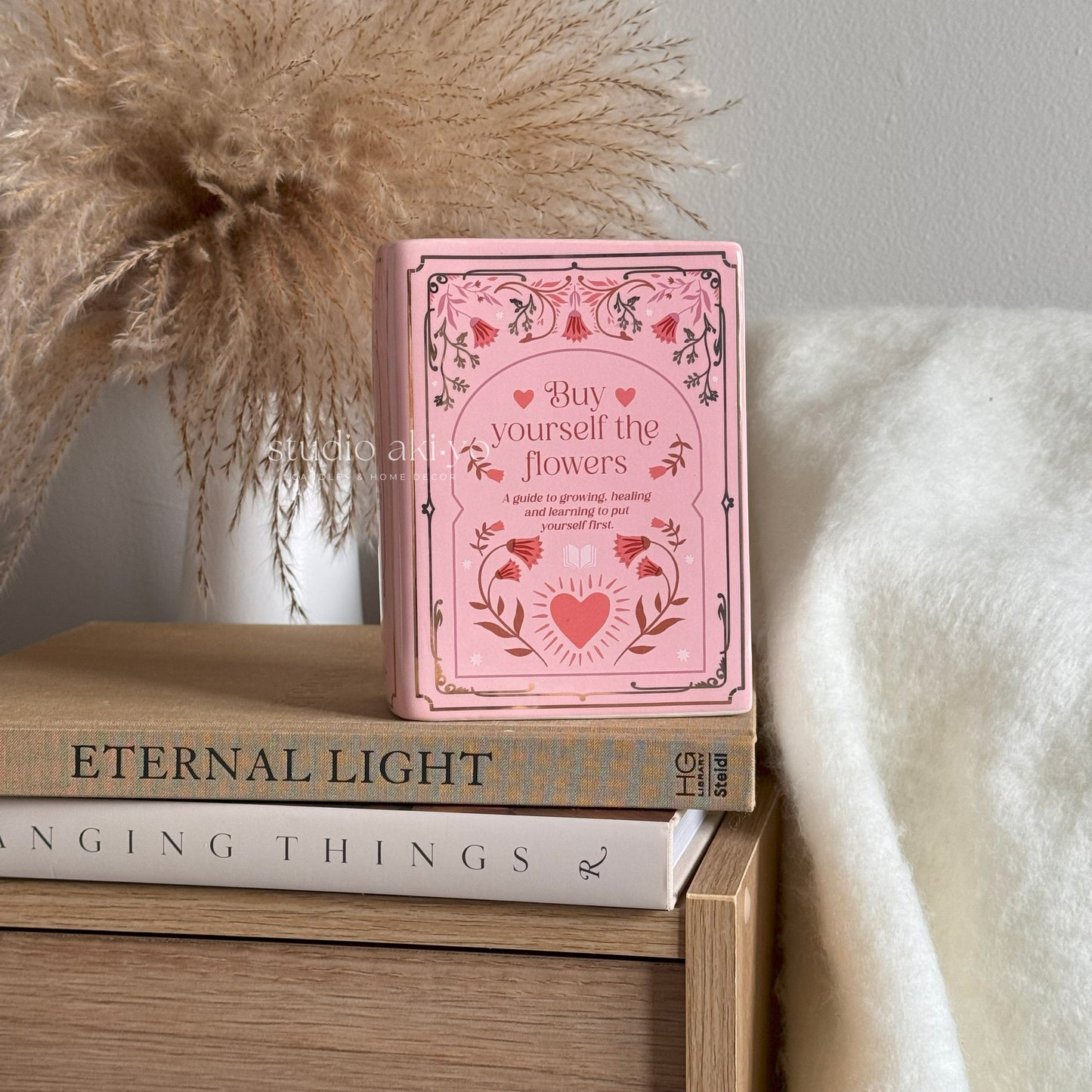Pink book titled 'Buy yourself the flowers' on a stack of books with decorative elements.