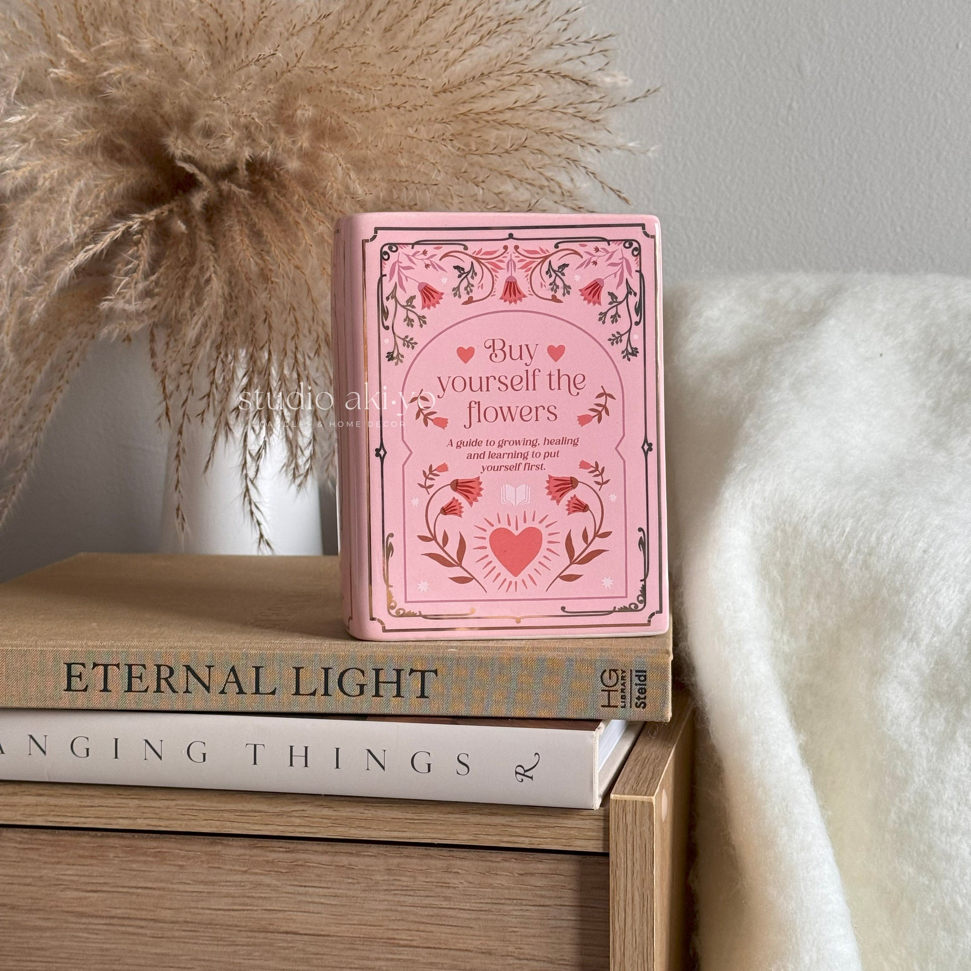 Pink book titled 'Buy yourself the flowers' on a stack of books with decorative elements.