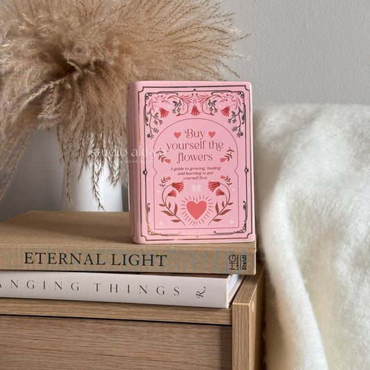 Pink book titled 'Buy yourself the flowers' on a stack of books with decorative elements.