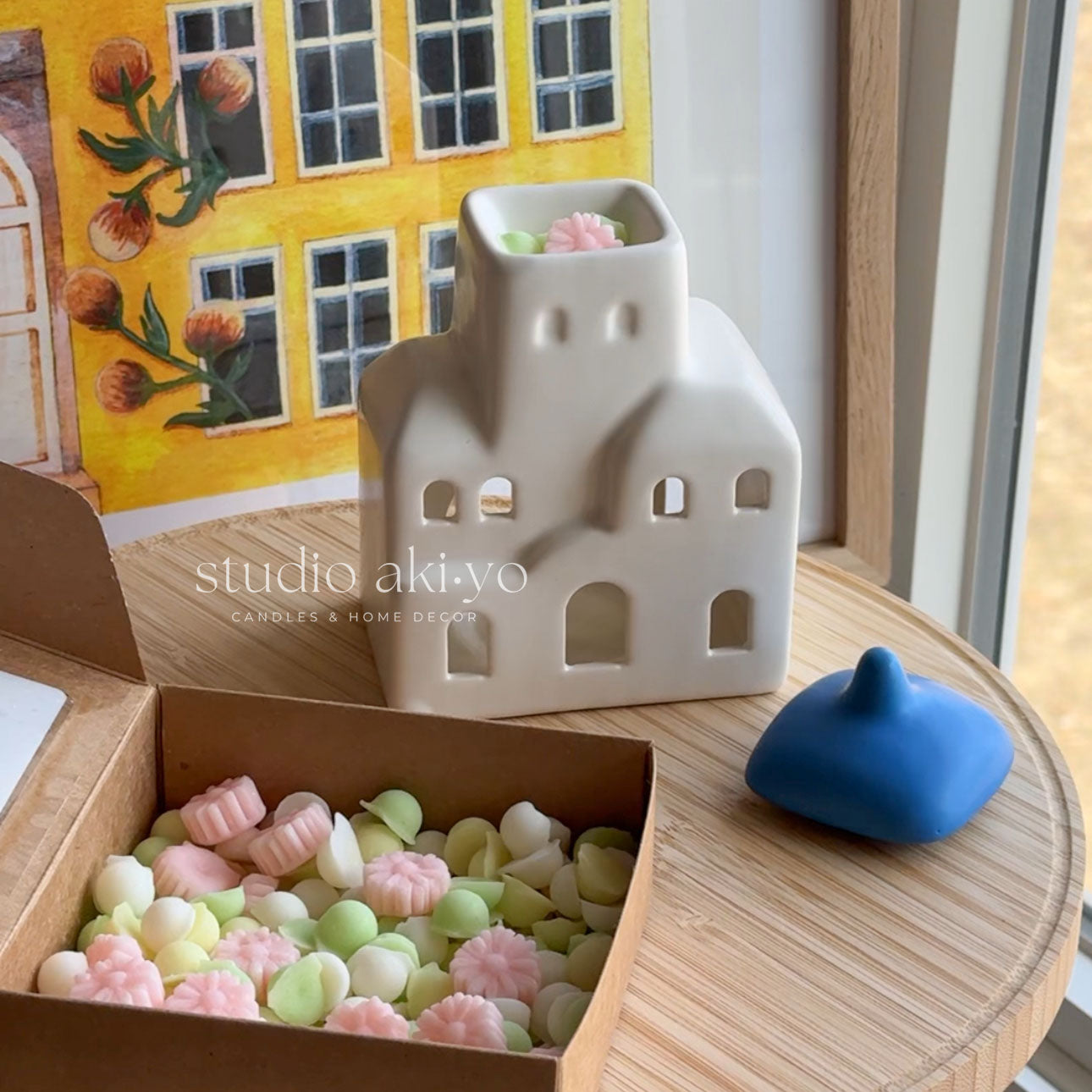 Ceramic house-shaped lantern with colorful beads in a box, next to a blue lid on a wooden surface.