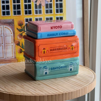 Stack of colorful boxes labeled 'Kyoto', 'Amalfi Coast', 'Marrakesh', and 'Santorini' on a wooden surface with a decorative background.