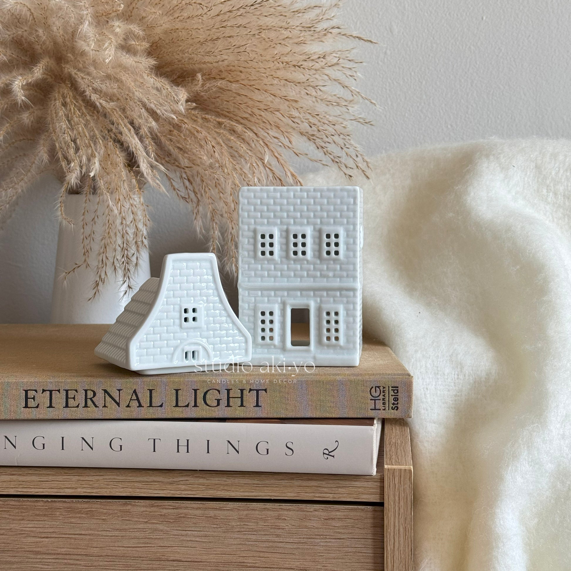 Decorative white ceramic houses on books with a pampas grass arrangement in the background.