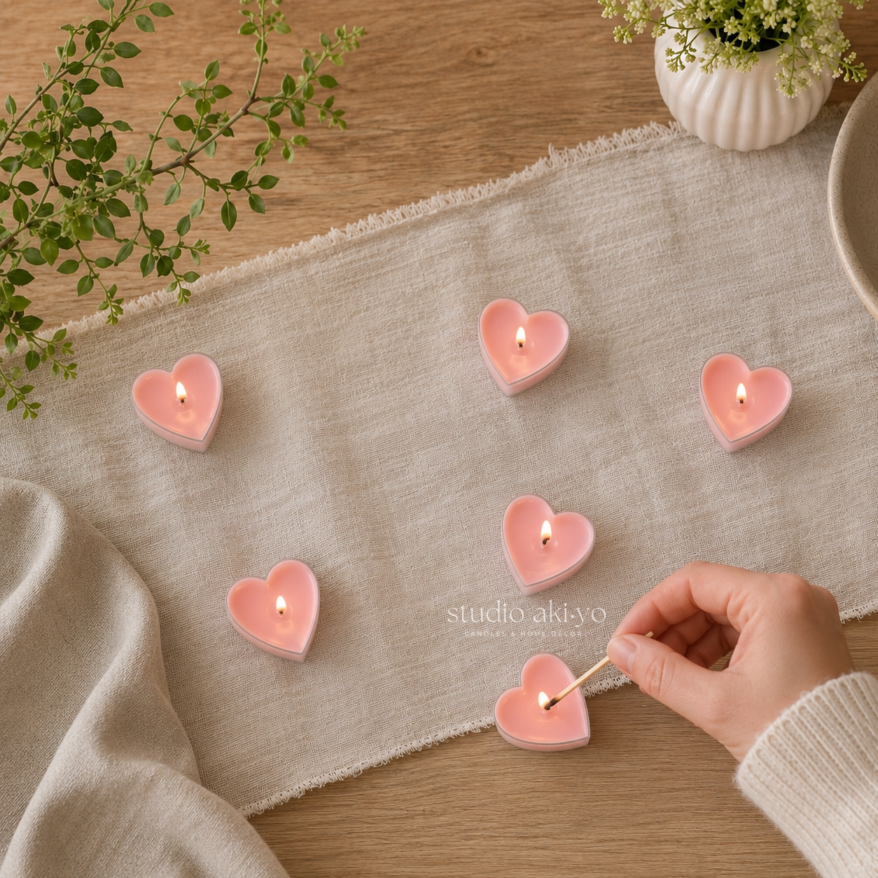 Heart-shaped candles being lit on a textured surface with a hand and match, surrounded by greenery.