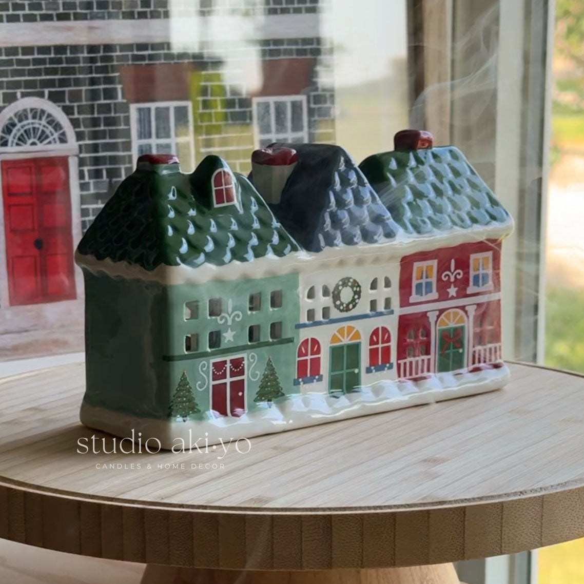 A ceramic Christmas village house candle holder with three colorful cottages under snowy roofs, displayed on a wooden stand in front of a window.