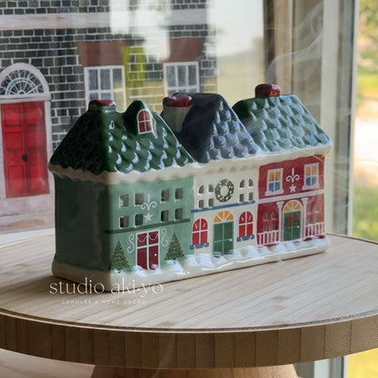 A ceramic Christmas village house candle holder with three colorful cottages under snowy roofs, displayed on a wooden stand in front of a window.