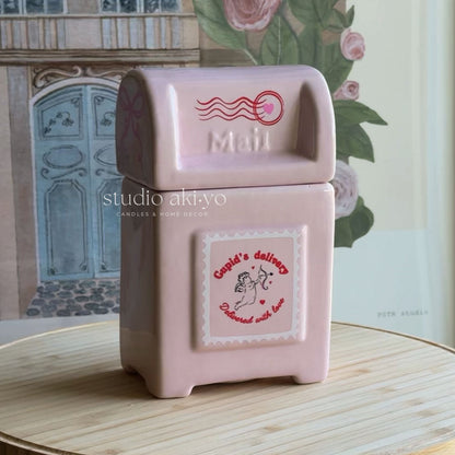 A pink ceramic mailbox candle holder with a vintage-style mail slot top and a front label reading 'Candi's delivery' and 'Discovered with love,' placed on a light wooden surface with a floral and architectural background.