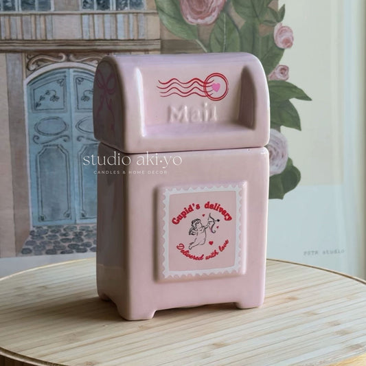 A pink ceramic mailbox candle holder with a vintage-style mail slot top and a front label reading 'Candi's delivery' and 'Discovered with love,' placed on a light wooden surface with a floral and architectural background.