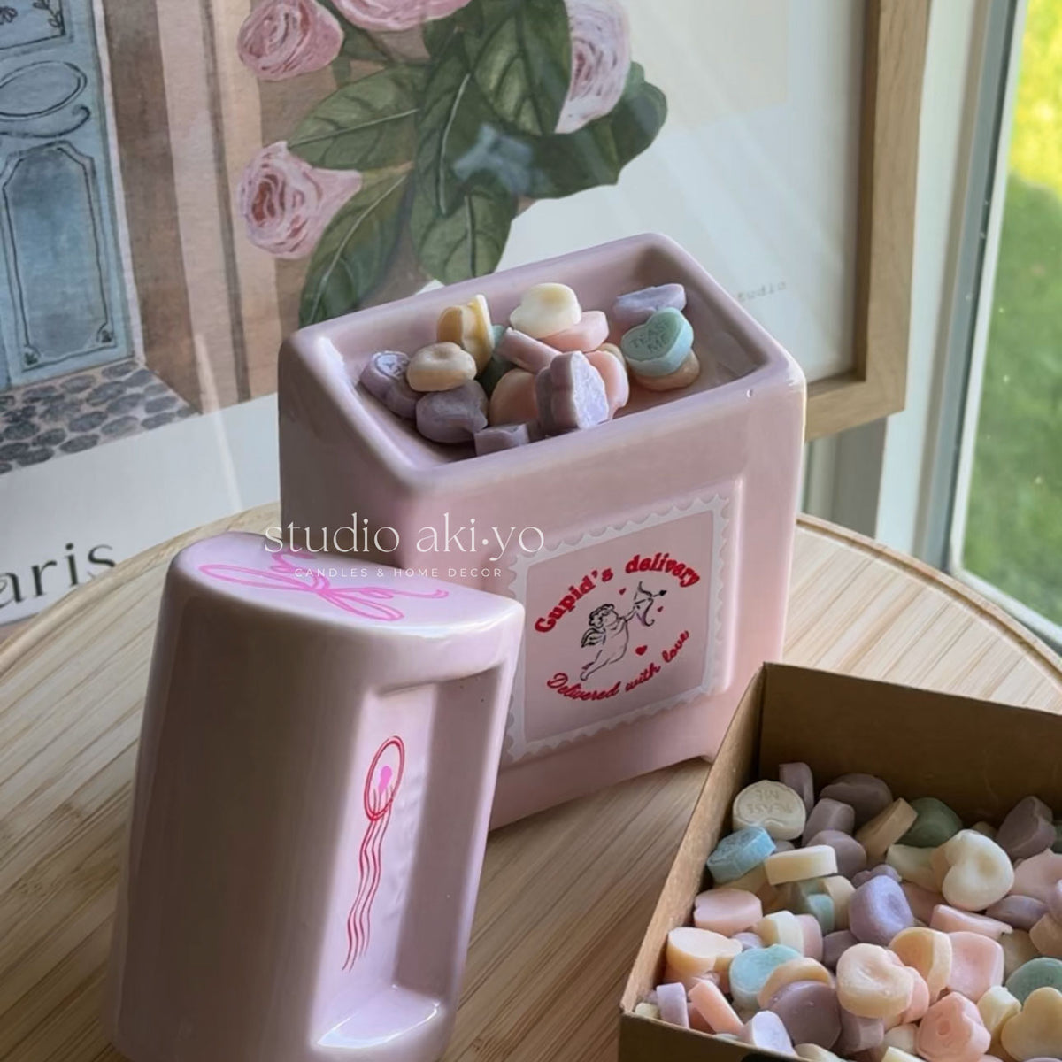 A pink ceramic candy dish set on a wooden table, with one dish open showing pastel candy pieces inside and another dish beside it. A cardboard tray of more pastel candies sits in the foreground.