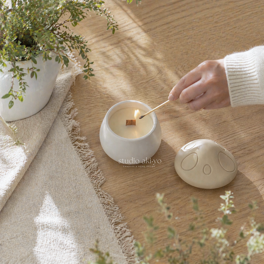 Person lighting a candle in a white container on a wooden surface with plants around.