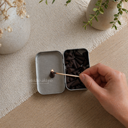 Hand lighting a match from a small metal box on a textured surface with plants in the background.