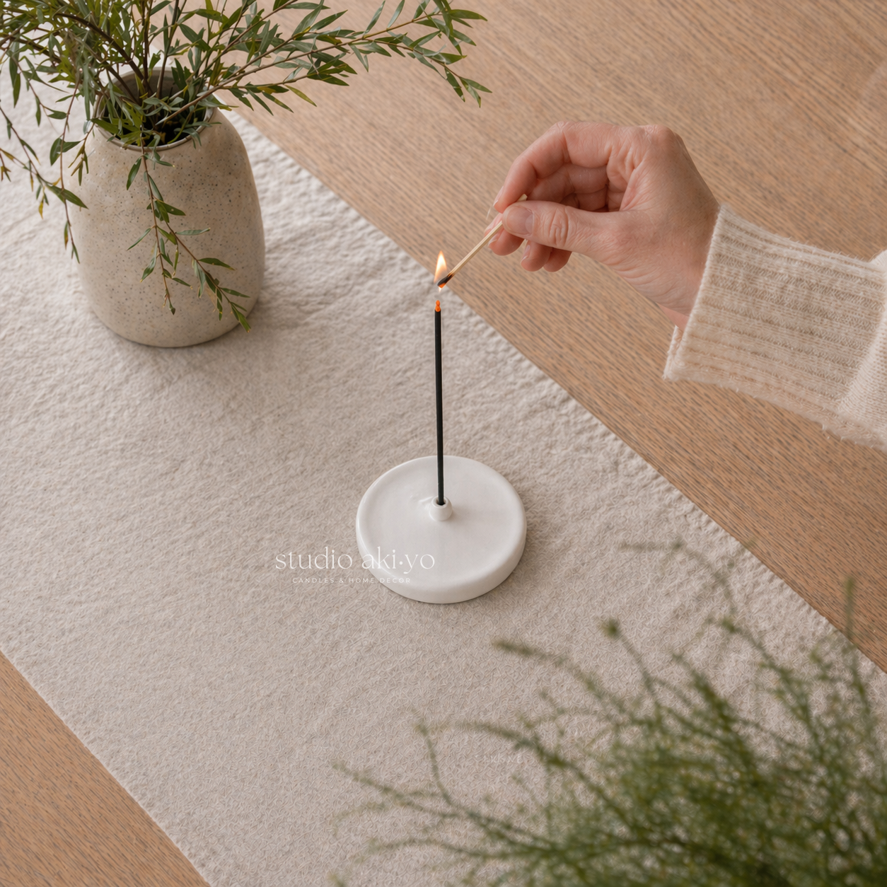 Person lighting an incense stick with a match on a marble base, next to a potted plant on a wooden surface.